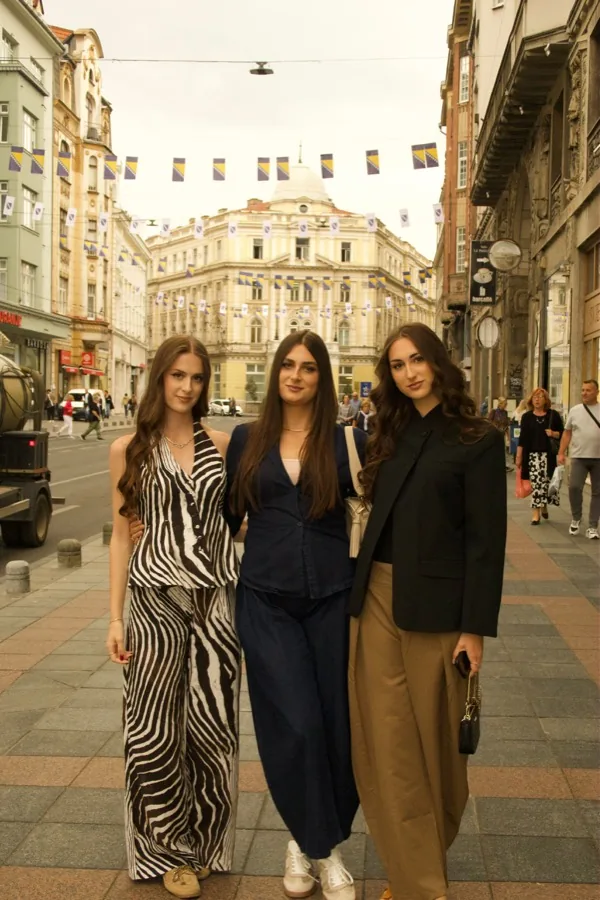 Three friends standing together on a Sarajevo street under strings of flags, the old post office behind them.