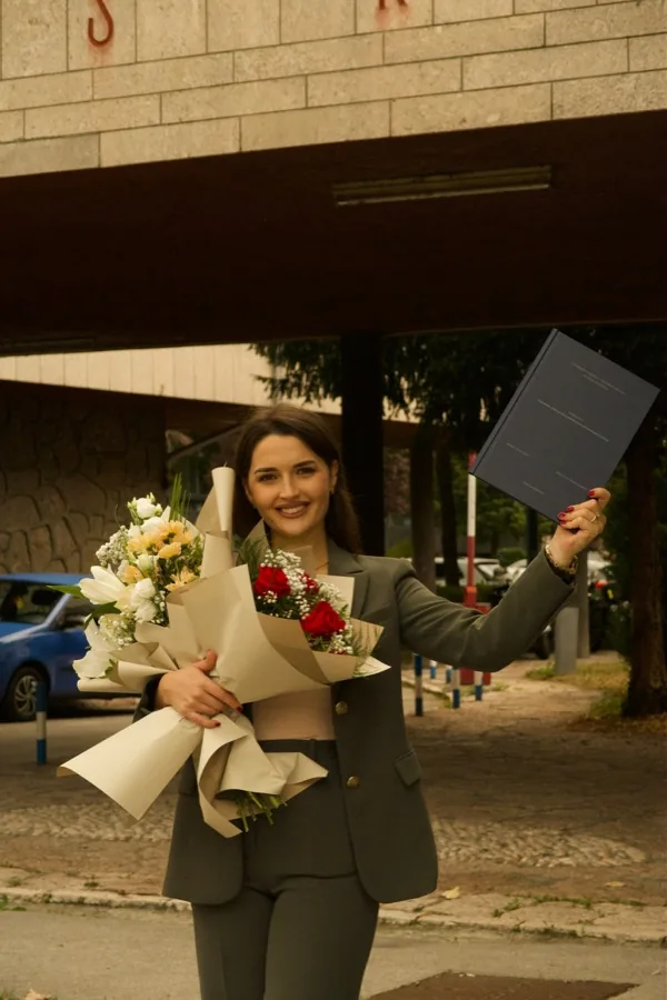 A graduate in a green trouser suit holding a large bouquet and raising her diploma folder outside the faculty.