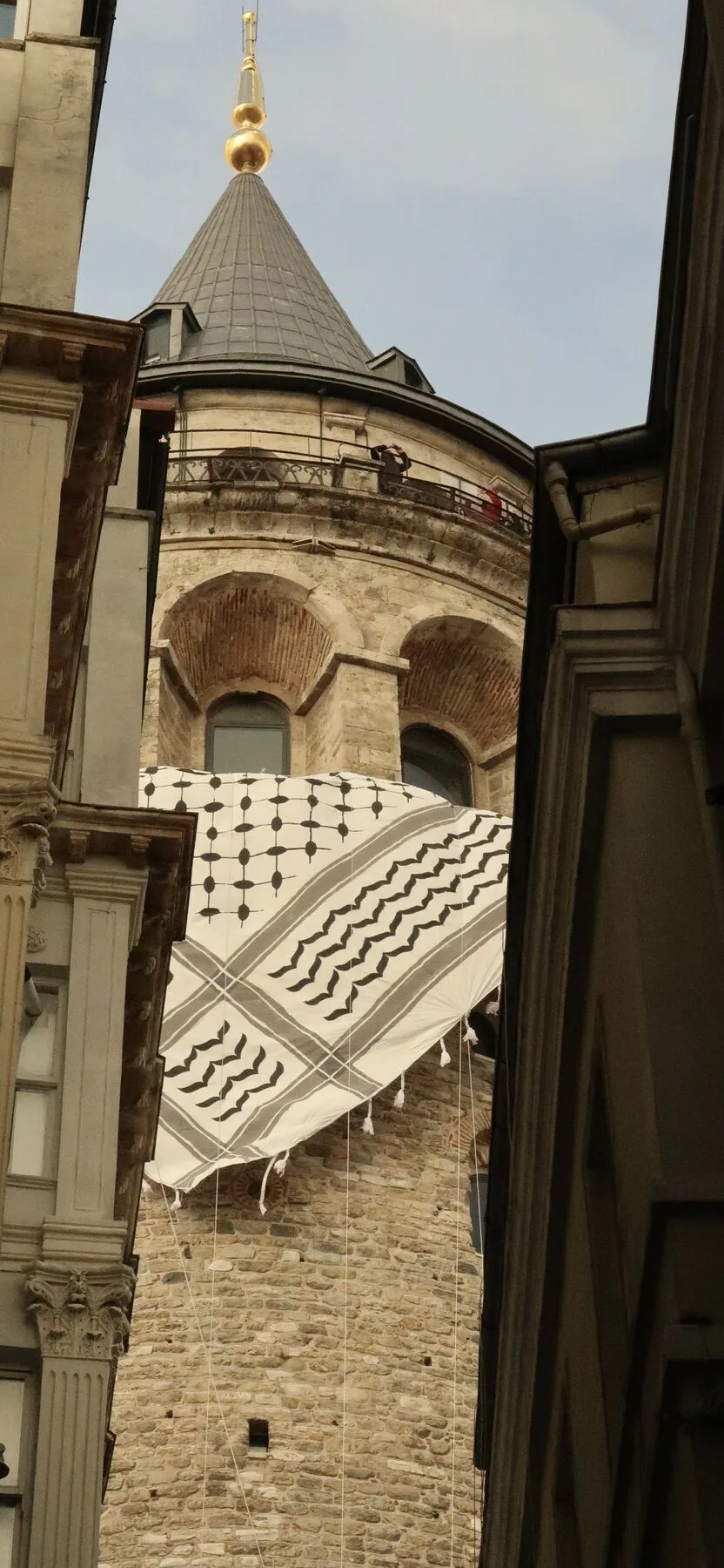 The Galata Tower seen through a narrow gap between buildings, a line of printed cotton hung across the street below it.