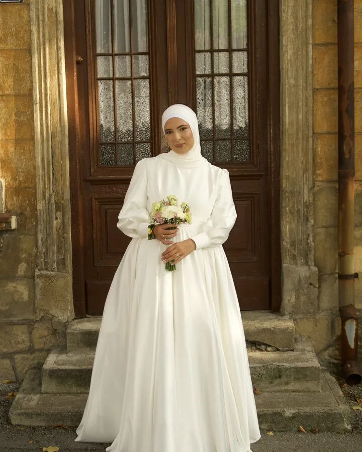 A bride in a white hijab and a long white gown standing on the steps of a weathered stone doorway, holding a small bouquet.