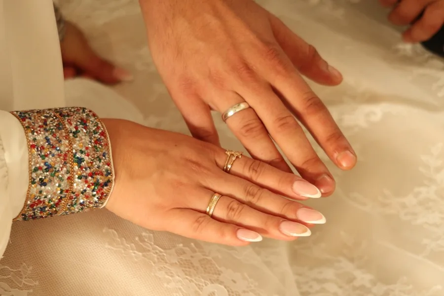 The couple's hands with wedding rings on white lace, her cuff embroidered in colour.