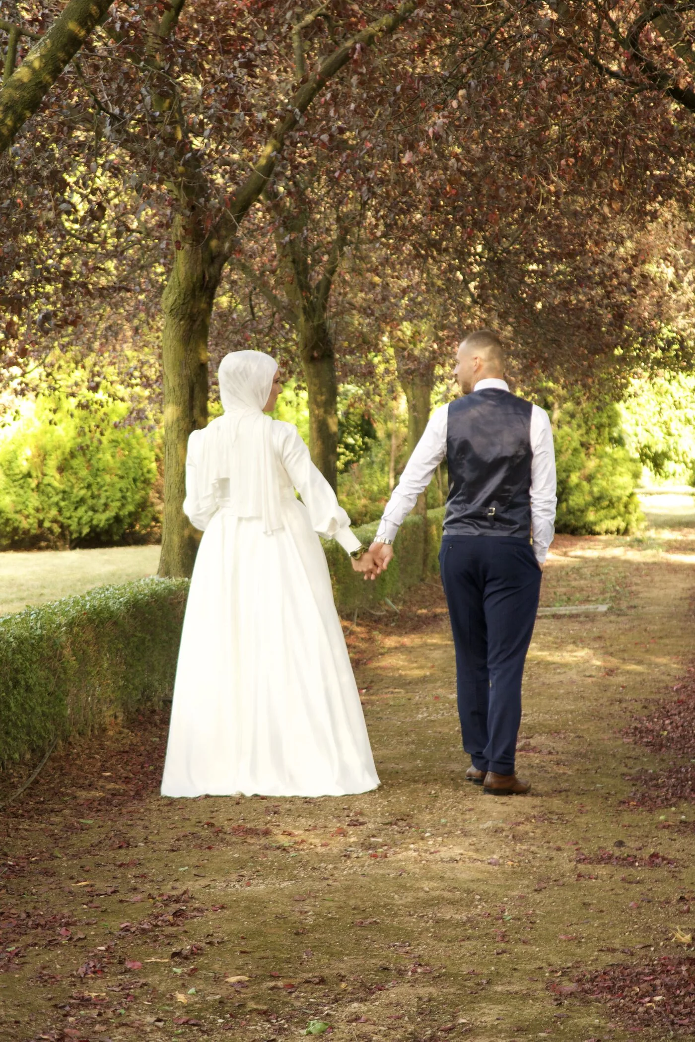 The bride and groom walking away hand in hand down a path under dark copper beech trees.